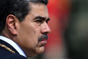 A imagem mostra Nicolás Maduro em Caracas, vestindo um terno azul com uma faixa colorida e insígnias. Ele está fazendo um gesto de punho cerrado, acompanhado por uma mulher que usa óculos escuros e um vestido branco. Ao fundo, há seguranças em trajes formais, e um fundo azul desfocado. O presidente da Venezuela, Nicolás Maduro